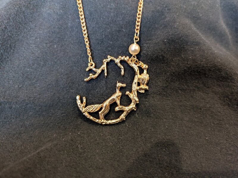 A gold necklace with a fox looking up at a crow. A pearl sits atop the tree.